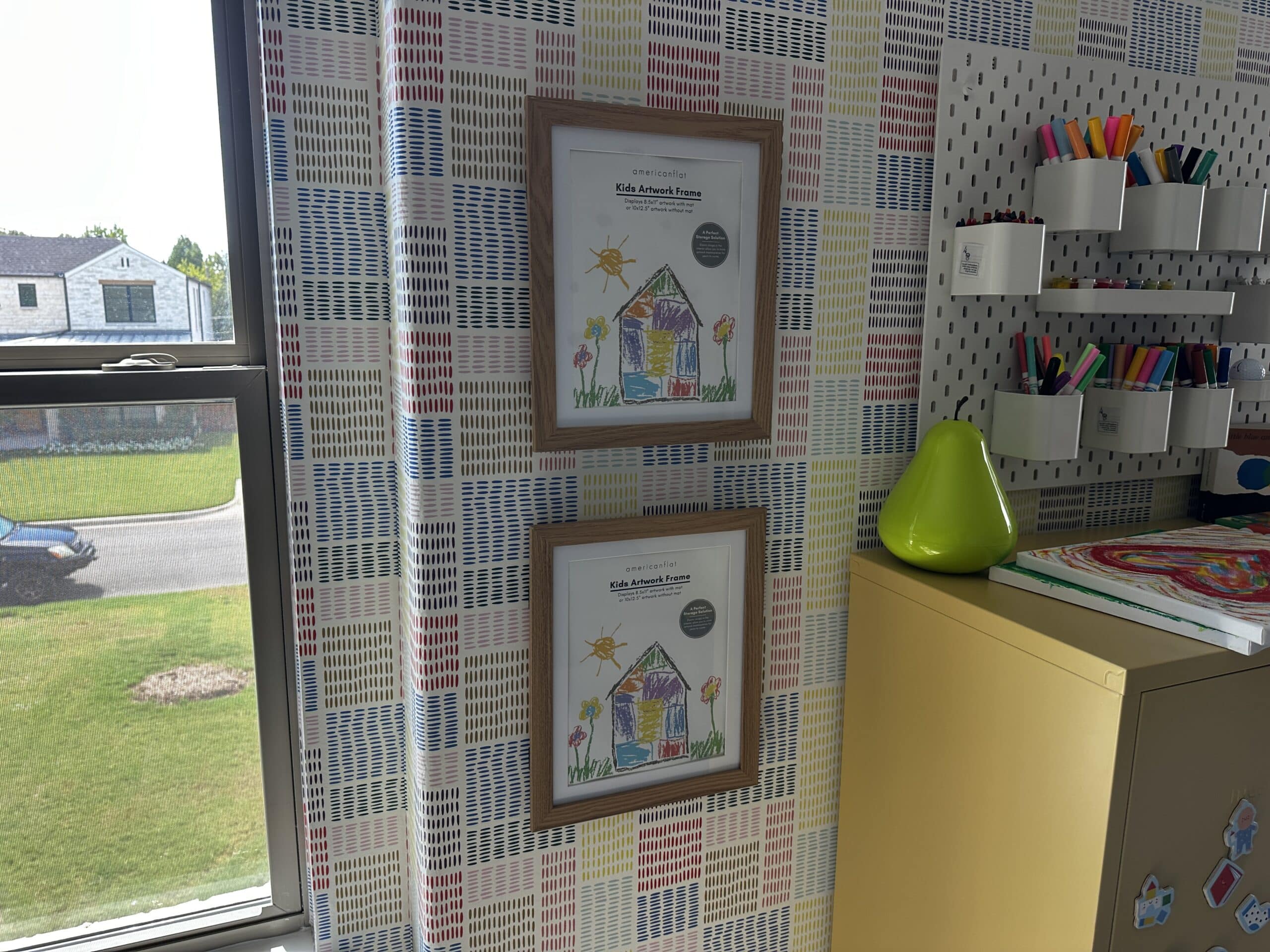 Two kids' artwork frames stacked one above the other on a patterned playroom wall