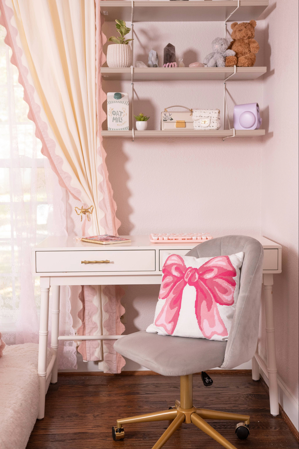 Three wall shelves above a white desk beside a window curtain with a brass bow tieback in a pink girls' bedroom